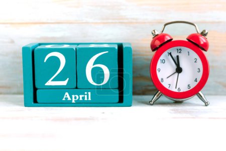 26. April. Blauer Würfelkalender mit Monat und Datum und roter Wecker auf Holzgrund