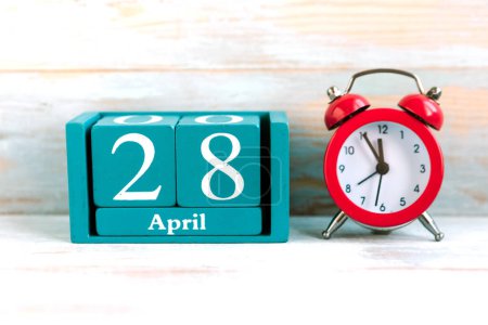 28 de abril. Calendario cubo azul con mes y fecha y despertador rojo sobre fondo de madera