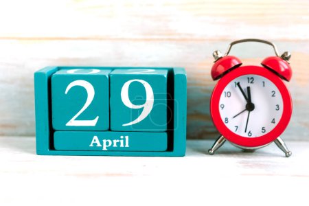29. April. Blauer Würfelkalender mit Monat und Datum und roter Wecker auf Holzgrund