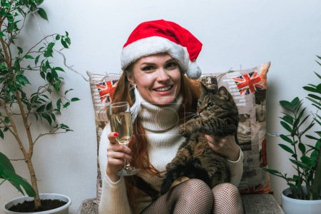 Porträt einer jungen rothaarigen Frau mit Weihnachtsmütze, Katze und Champagnerglas im Wohnbereich