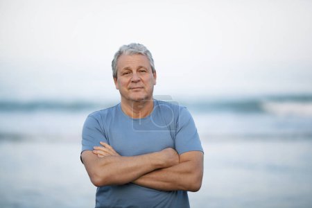 Middle shot of a senior man in blue t-shirt with arms crossed on the chest. Person looking to the camera with a light smile. View against blurry background of sea and sky