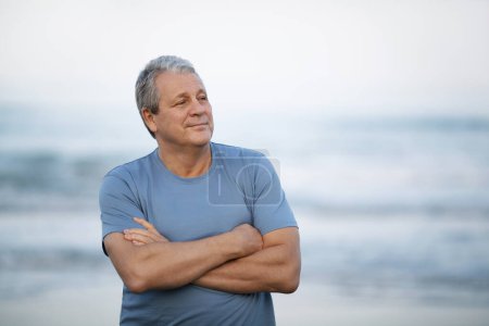 Foto de Middle shot of a smiling senior man looking away with arms crossed on the chest. Cheerful person on blurry background of sea and sky - Imagen libre de derechos