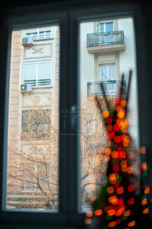 Foto de Inside view of a window in the apartment overlooking the neighbouring house and bare tree. Decoration item with blurry red lights in foreground. Focus on the opposite building - Imagen libre de derechos