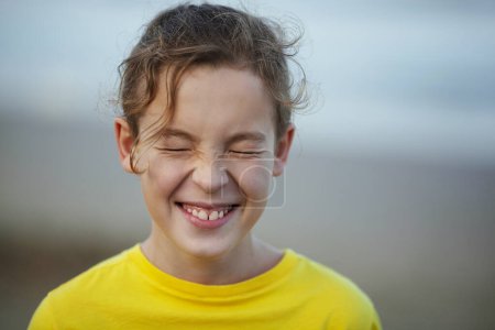 Foto de Close-up shot of a boy laughing with eyes closed tight. Happy child with curly hair in yellow t-shirt. Outdoor view against blurry background. Expecting a surprise - Imagen libre de derechos
