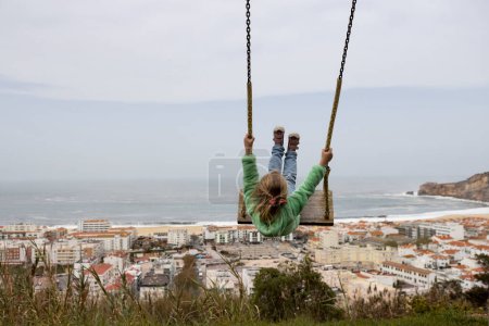 Foto de Back view of a kid on the swing. Child enjoying swinging and looking at ocean scene with city along the coast. Traveling to Nazare, Portugal - Imagen libre de derechos