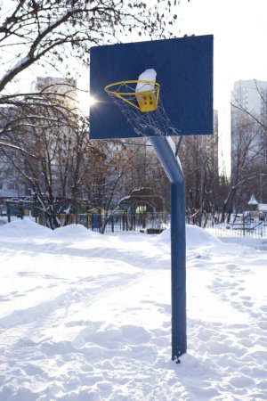 Foto de The wind shakes the basketball net. Blue basketball backboard with snow. Winter, buildings, playground and trees are in the background - Imagen libre de derechos