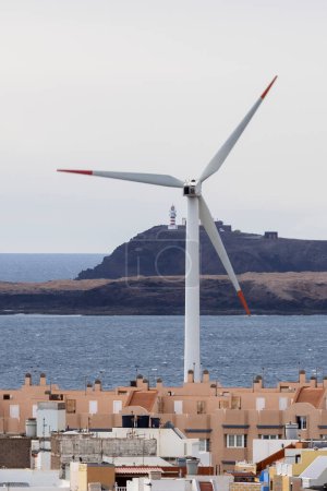 Foto de Turbina eólica ubicada en una pequeña ciudad cerca del mar azul ondulante contra el faro en la colina - Imagen libre de derechos