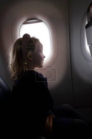 Foto de Vista lateral de la niña preadolescente sonriente en vestido sentado en el asiento del pasajero en el avión cerca de ojo de buey - Imagen libre de derechos