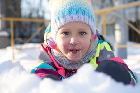 Foto de Retrato de linda chica sonriente en colorido sombrero de punto y bufanda tumbada en la nieve y mirando a la cámara - Imagen libre de derechos