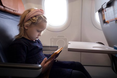 Foto de Vista lateral de la niña preadolescente enfocada en el vestido sentado en el asiento en el avión y viendo dibujos animados a través de la tableta - Imagen libre de derechos