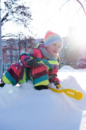 Foto de Cuerpo completo de niña preadolescente positiva en ropa caliente colorida sentada en la colina nevada con juguete para hacer bolas de nieve mientras juega en el patio - Imagen libre de derechos