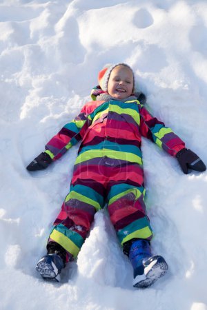 Foto de Desde arriba todo el cuerpo de la niña preadolescente riendo en ropa interior colorida haciendo ángel de nieve mientras juega al aire libre en el día de invierno soleado - Imagen libre de derechos