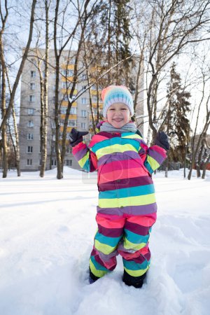 Foto de Chica alegre en ropa exterior de colores brillantes y sombrero de pie en la carretera nevada en el patio cerca de edificio residencial - Imagen libre de derechos