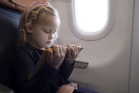 Foto de Chica preadolescente concentrada con el pelo rubio usando diadema sentado en el asiento del pasajero en el avión y viendo dibujos animados a través de la tableta - Imagen libre de derechos