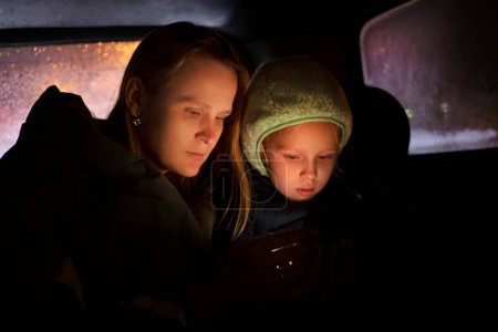 Foto de Mujer seria en ropa de abrigo sentado en el coche con su hija pequeña en el sombrero y el uso de teléfono inteligente durante el viaje nocturno - Imagen libre de derechos