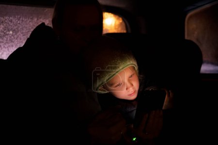 Foto de Chica positiva en el sombrero sentado en el asiento trasero del coche y abrazarse con la madre mientras se utiliza el teléfono inteligente durante el viaje nocturno - Imagen libre de derechos