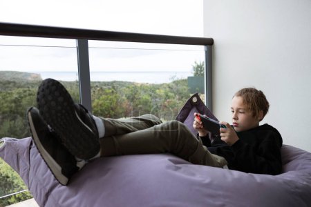 Foto de Adolescente disfrutando de su tiempo libre y jugando en la consola de mano. Niño tumbado en la cama beanbag en el balcón con vista a la naturaleza - Imagen libre de derechos