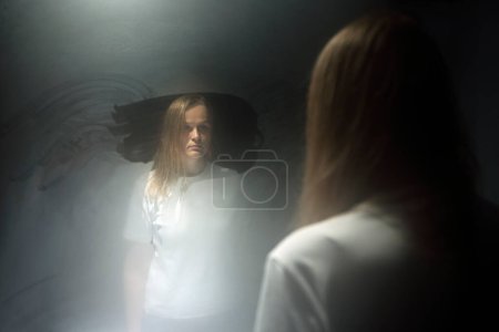 Foto de Mujer rubia en el reflejo del espejo sudoroso. Mujer con camiseta blanca mirándose fijamente con la mirada en blanco. Crisis de edad - Imagen libre de derechos
