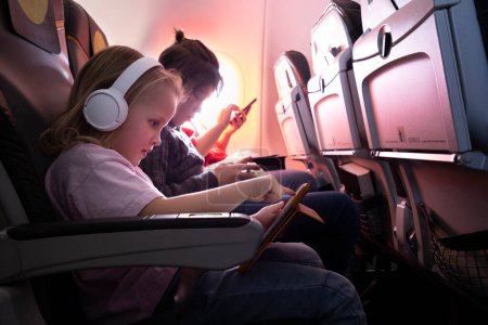 Foto de Viaje familiar en avión. Niña en auriculares inalámbricos usando tableta digital, niño y mujer entretenidos con teléfonos - Imagen libre de derechos