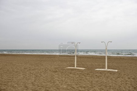 Foto de Playa de arena vacía con duchas. Vista al mar con cielo opaco, la temporada de vacaciones ha terminado - Imagen libre de derechos