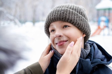 Foto de Sonriente niño en ropa de invierno al aire libre y las manos de la madre aplicando crema a su cara para proteger la piel del viento y el frío - Imagen libre de derechos