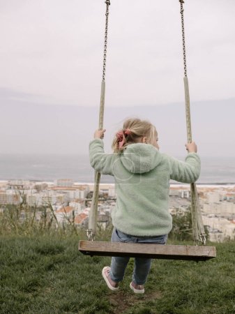 Foto de Una niña de 5 años se balancea sobre simples columpios de madera en la cima de una colina, vistos desde atrás. En la distancia, una ciudad y el océano son visibles - Imagen libre de derechos