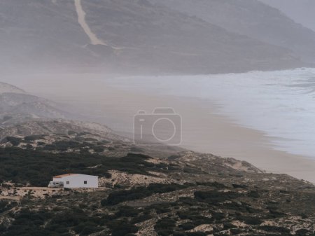 Foto de Una escena serena y tranquila con una casa solitaria a orillas del Océano Atlántico en Portugal. Los colores tenues y la atmósfera brumosa creada por las olas que se estrellan proporcionan una sensación - Imagen libre de derechos