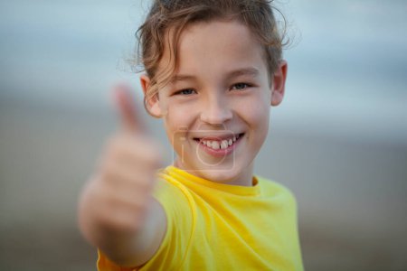 Foto de Close-up shot of a happy boy showing thumbs-up. Child with a broad smile in yellow t-shirt on blurry background. Positive thinking - Imagen libre de derechos