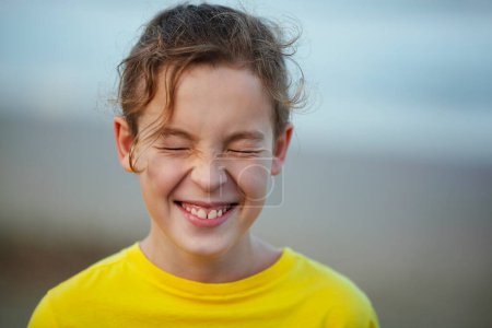 Foto de Close-up shot of a boy laughing with eyes closed tight. Happy child with curly hair in yellow t-shirt. Outdoor view against blurry background. Expecting a surprise - Imagen libre de derechos