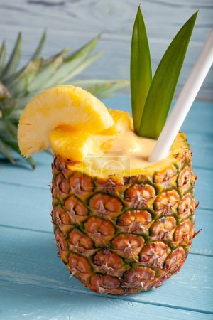 Photo for A vertical photo of a pina colada cocktail on a blue wooden background with pineapple pieces, a white straw, two green leaves, and a sliced pineapple crown lying in the blurred background - Royalty Free Image