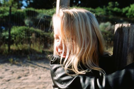 Foto de Sonriente chica de pelo rubio en chaqueta de cuero negro mirando hacia otro lado mientras trepa cerca de madera vieja en el campo - Imagen libre de derechos