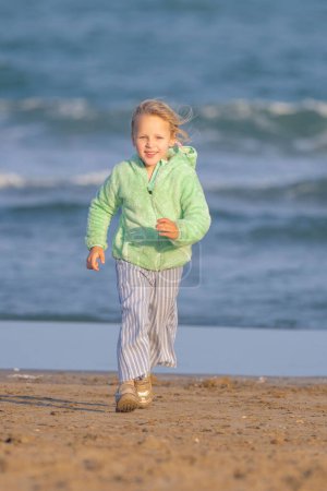Foto de Largo tiro - foto vertical de una niña de 5 años, rubia, con una chaqueta verde recorre la playa junto al mar, con una sonrisa - Imagen libre de derechos