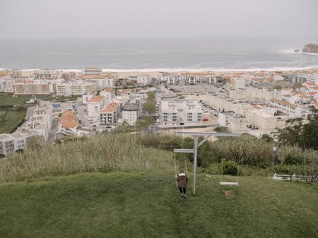 Foto de Un adolescente de 12 años con una chaqueta disparada por la espalda se balancea en un columpio situado en una colina con vistas a la ciudad y al océano, Portugal - Imagen libre de derechos