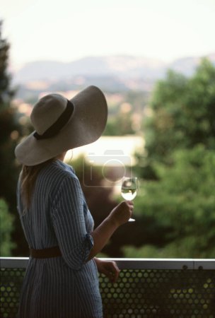 Foto de Una mujer, por detrás, con un vestido y un sombrero, se para con una copa de vino en la mano - Imagen libre de derechos