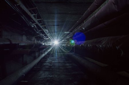 Dark underground concrete utility communication tunnel with pipes and wires in cold colors.