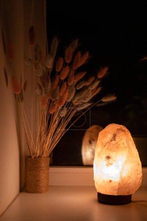 Photo for Warm light from a salt lamp illuminating a bouquet of lagurus in the evening. Warm light. Room lighting. Home comfort. The benefits of a salt lamp. Dry flowers in the interior. Apartment decoration. - Royalty Free Image