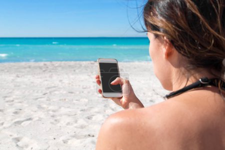 Foto de Mujer de vacaciones en la playa sosteniendo el teléfono inteligente, mensajes de texto y tocando. Vacaciones de verano con el mar azul en el fondo - Imagen libre de derechos