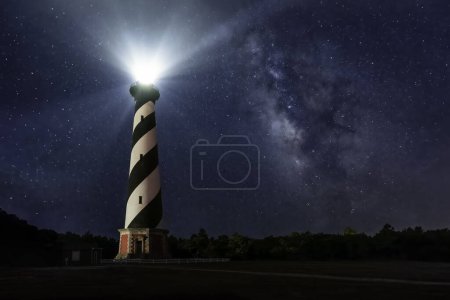 Téléchargez les photos : Le phare le plus haut d'Amérique, The Cape Hatteras Light on the Outer Banks of North Carolina, brille dans le ciel étoilé de la nuit d'été avec le centre galactique de la Voie lactée visible. - en image libre de droit