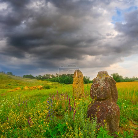 old  ancient stone statue among green fields under dense cloudy sky, open air museum scene