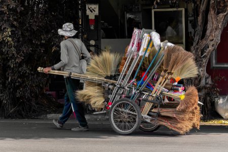 Téléchargez les photos : Bangkok, Thaïlande, 18 janvier 2023 : La vie quotidienne d'un colporteur avec son chariot à deux roues tiré vendant des outils de nettoyage - brosses à récurer, balais, balayeuses et vadrouilles. - en image libre de droit