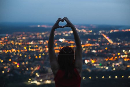 Téléchargez les photos : La fille montre son cœur avec ses mains sur le fond des lumières de la ville du soir. - en image libre de droit