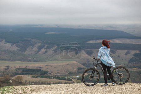 Téléchargez les photos : Une femme au sommet d'une montagne avec un vélo regarde le panorama de montagne admiratif lors d'une balade à vélo - en image libre de droit