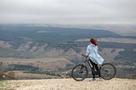 Foto de Una mujer en la cima de una montaña con una bicicleta mira el panorama de la montaña durante un viaje en bicicleta - Imagen libre de derechos