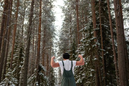 Téléchargez les photos : Jeune dans un t-shirt homme à l'apparence athlétique dans la forêt d'hiver entouré de flocons de neige. Concept sport et acclimatation au froid. Photo de haute qualité - en image libre de droit