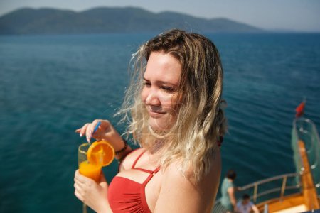 Photo for Happy busty woman on yacht. Lady in girl in coral bikini holding orange fresh drink. Cool drink and fresh aquamarine sea. Living a happy life. - Royalty Free Image