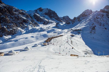 Photo for Shymbulak mountain resorts ski slopee with eco hotel, yurts, skiers and snowboarders. Skiers at slopes of ski resort Shymbulak. - Royalty Free Image