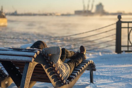 Téléchargez les photos : Belle femme portant des vêtements d'hiver relaxant sur le transat sur le coucher de soleil givré d'hiver - en image libre de droit