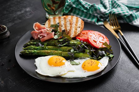 Foto de Breakfast. Fried egg, bread toast, green asparagus, tomatoes and jamon on black plate. - Imagen libre de derechos