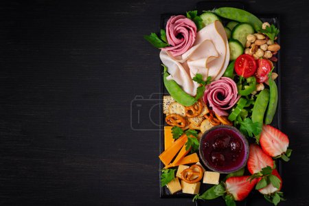 Photo for Antipasto platter cold sliced ham, salami, crackers, strawberries, vegetables and cheese platter on  board over dark background. Appetizers table with italian antipasti snacks. Top view - Royalty Free Image