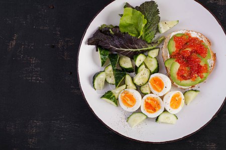 Téléchargez les photos : Breakfast. Healthy open sandwich on  toast with avocado and red caviar, boiled eggs, cucumber salad on white plate. Healthy protein food. Top view - en image libre de droit